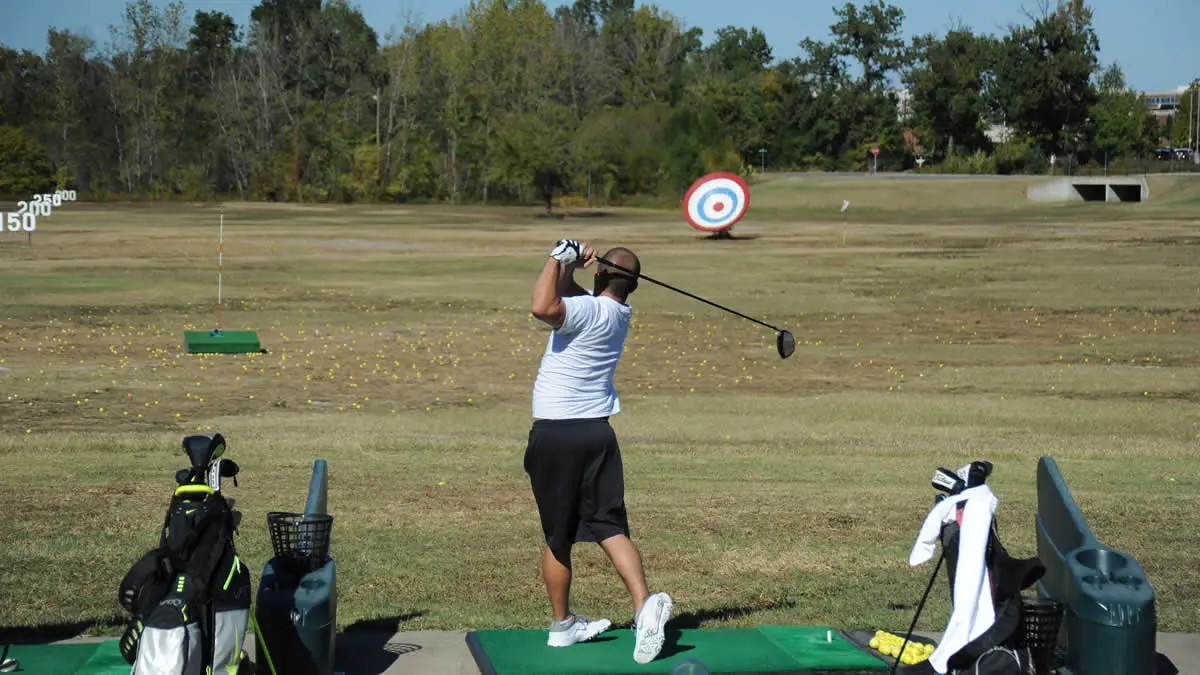 View behind golfer as they take a swing on a driving range tee line with driving range yardage markers in the field and a bullseye satellite dish